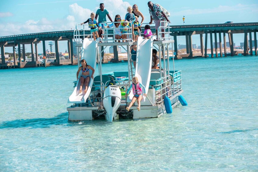 A group of people on a boat with a water slide, a pier in the background, and clear blue water. 3