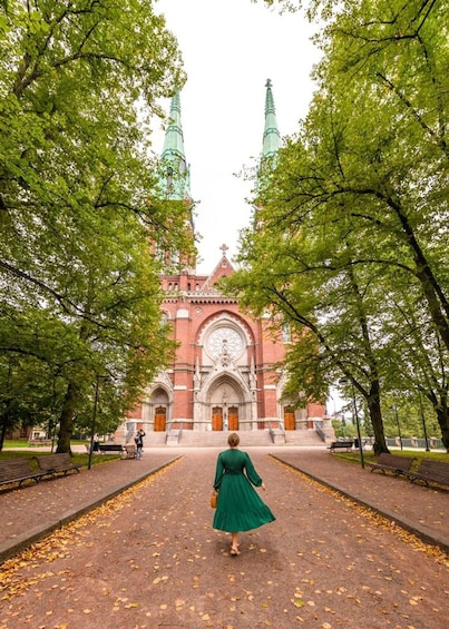 A woman in a green dress walking on a leaf-covered path towards a historic church with two tall spires. 1