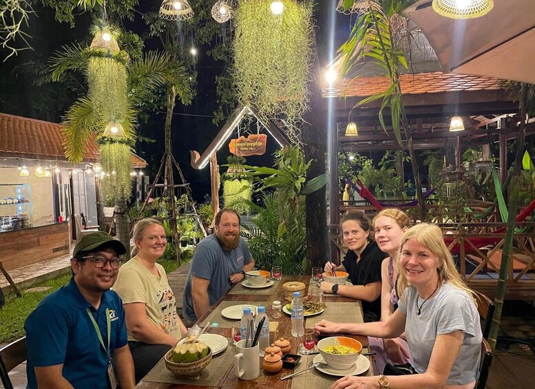 Ryhmä ihmisiä syömässä ulkoilmarestaurangissa, jossa on roikkuvia kasveja ja perinteinen sisustus. 1