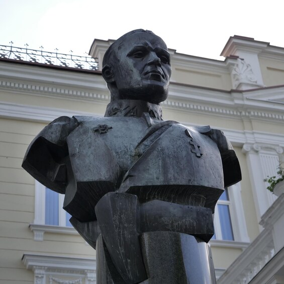 Una estatua de un hombre con uniforme militar y medallas, frente a un edificio de arquitectura clásica. 2