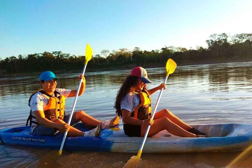 Two people in a blue kayak on a calm river, each holding a yellow paddle. 1