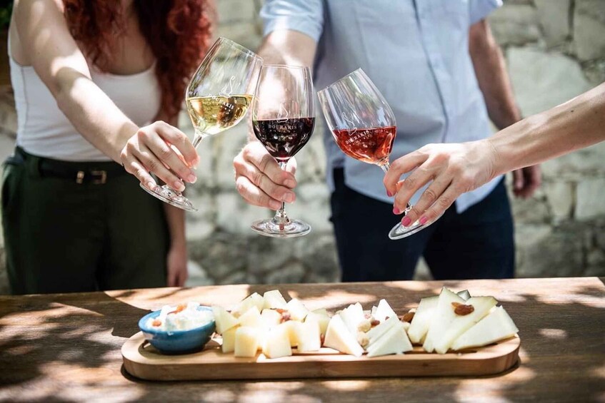 Three people toasting with wine glasses, a wooden table with cheese and a bowl of nuts. 3