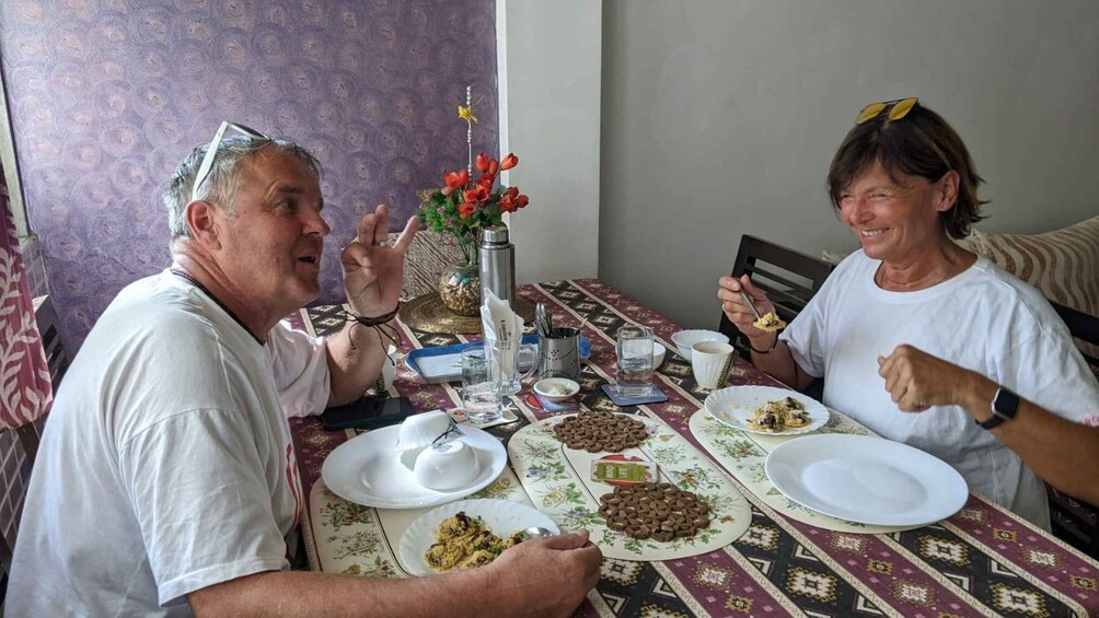 Two people sitting at a table with plates of food, a thermos, and a vase with flowers. 2
