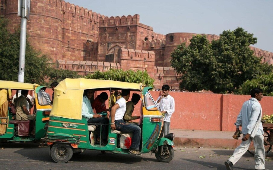 An auto-rickshaw on a road with passengers, a man walking, and a large red fort in the background. 1