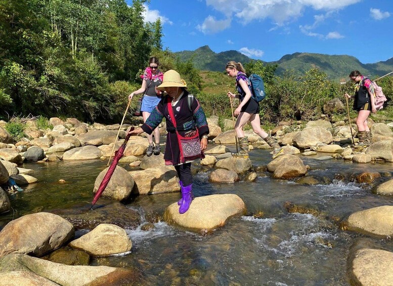 People crossing a rocky stream in a forested area with mountains in the background. 5