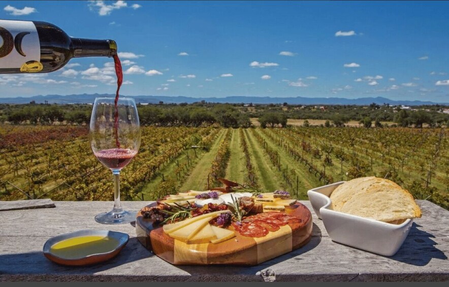 A wine bottle pouring into a glass, a cheese board with various cheeses, a bowl of bread, and a small dish of olive oil, set against a vineyard backdrop. 3