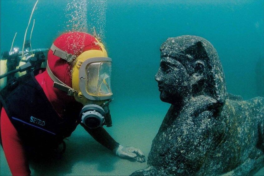 A diver in red and black gear with a yellow helmet and mask, interacting with a stone statue of a lioness. 1