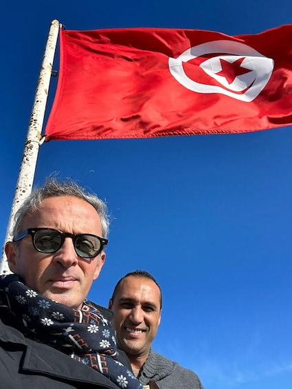 Dos personas de pie frente a un asta con una bandera roja que muestra una estrella blanca y una luna creciente. 1