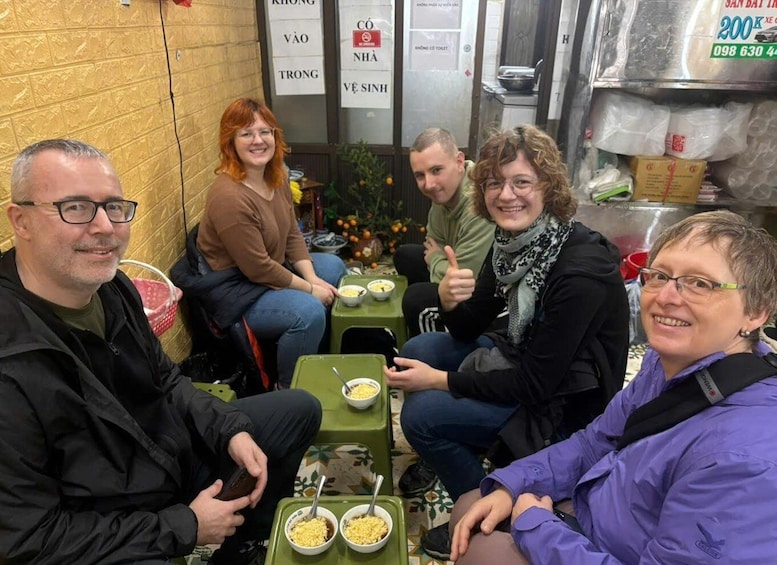 A group of people sitting at a table with bowls of food, in a room with a tiled wall and a sign with Vietnamese text. 2