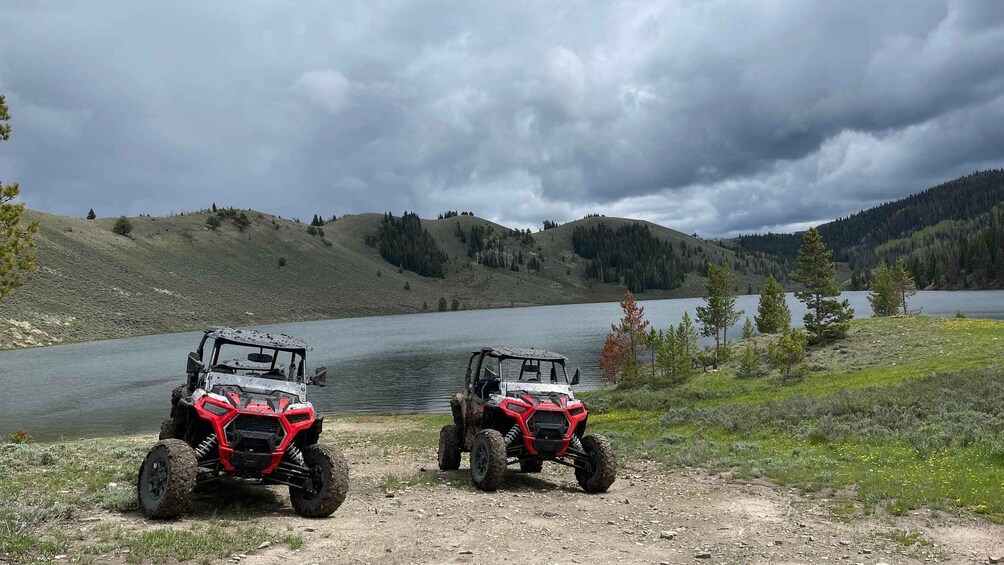 Two red off-road vehicles parked on a dirt path by a lake with hills in the background. 3