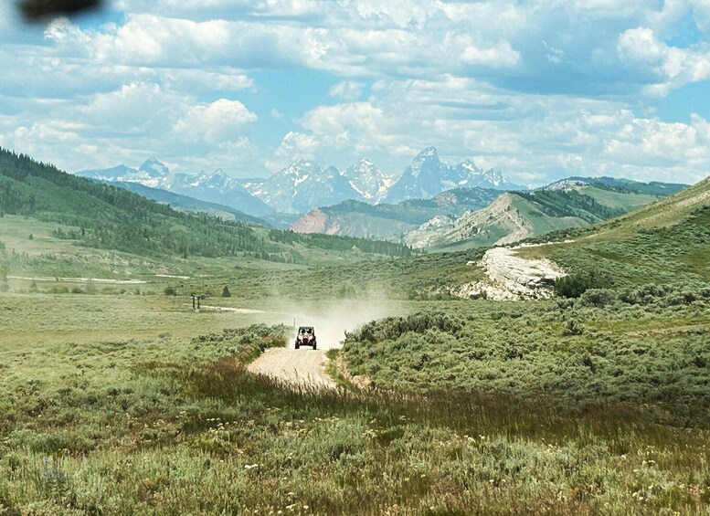 A jeep driving on a dirt road in a mountainous area with green fields and snow-capped peaks in the background. 1