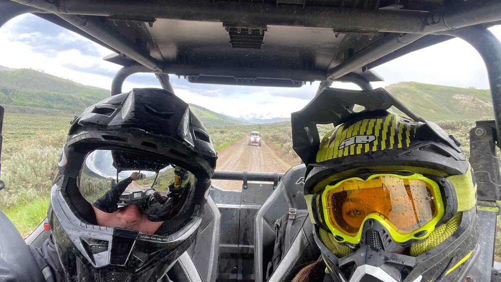 Two people in a vehicle with helmets, a road, and a hilly landscape. 2