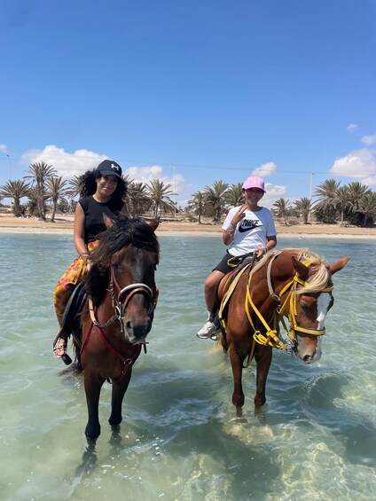 Two people riding horses in shallow water near a beach with palm trees. 3