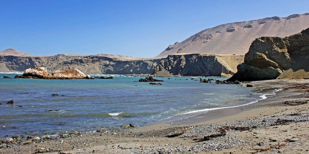 A rocky beach with clear water, a large rock formation, and a mountain in the background. 1