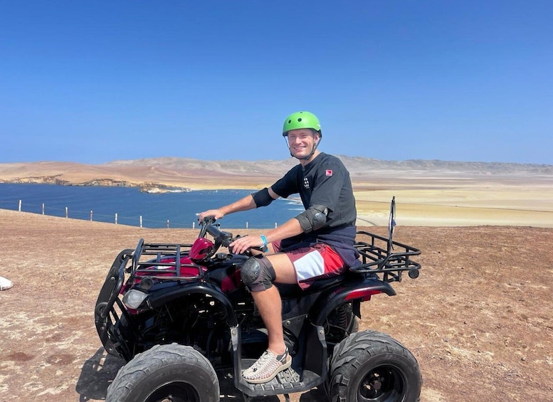 A person on a quad bike with a green helmet, sitting on a dirt path with a beach and hills in the background. 1