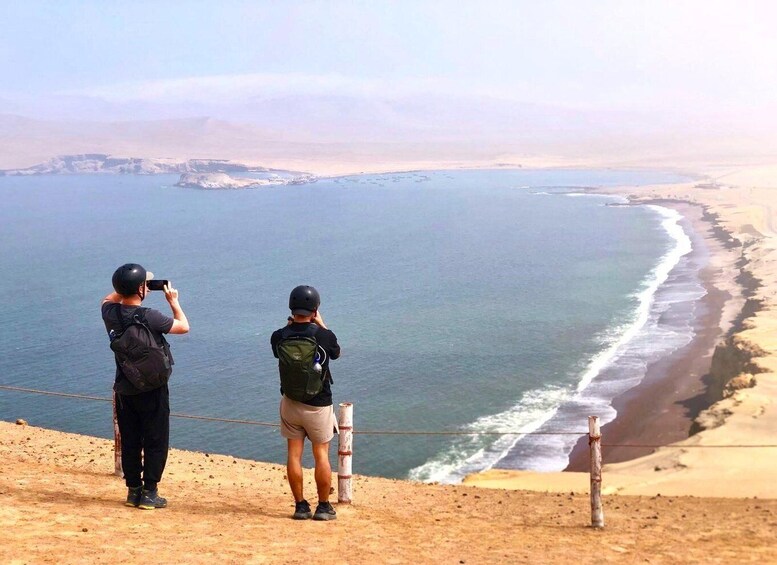 Two people taking photographs of a coastal landscape with a sandy beach and a rocky outcrop in the distance. 3