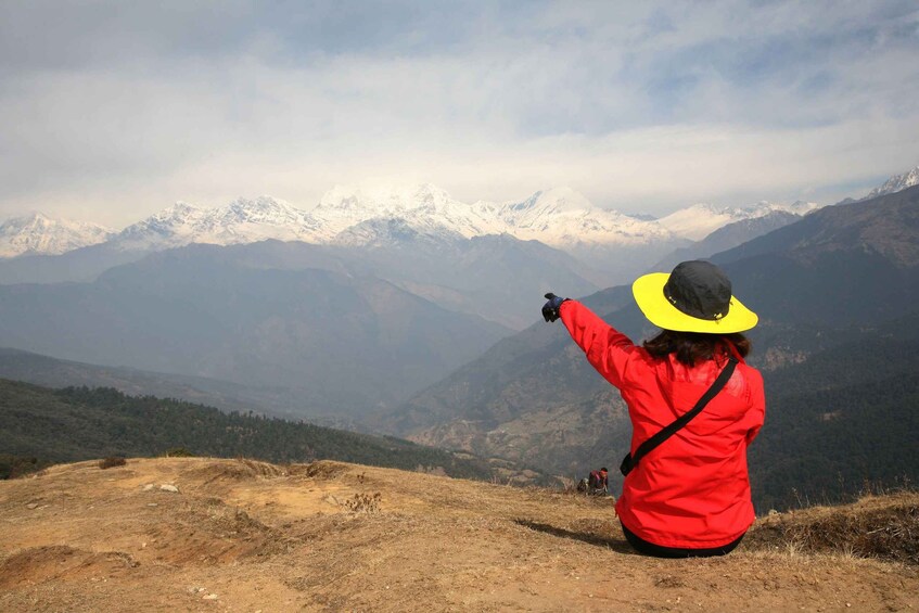 Una persona con chaqueta roja y sombrero amarillo sentada en una colina, señalando hacia una cordillera con picos nevados. 4