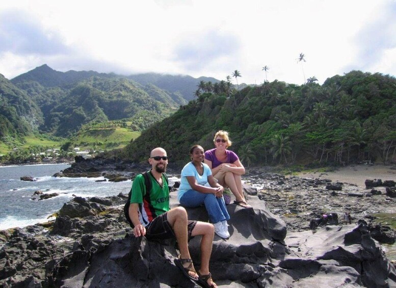 Three people sitting on rocks by a coastal area with mountains and palm trees in the background. 3