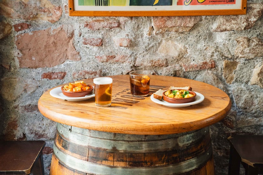 A wooden table with two bowls of food and two glasses of beer, set against a stone wall. 2