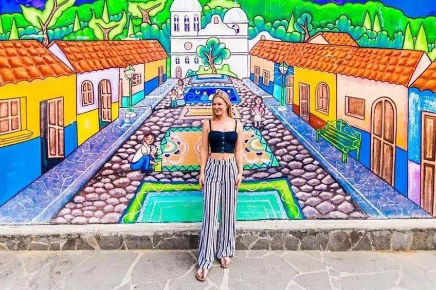 Une personne se tient devant une fresque colorée représentant une scène de rue avec des bâtiments, une fontaine et des arbres. 1
