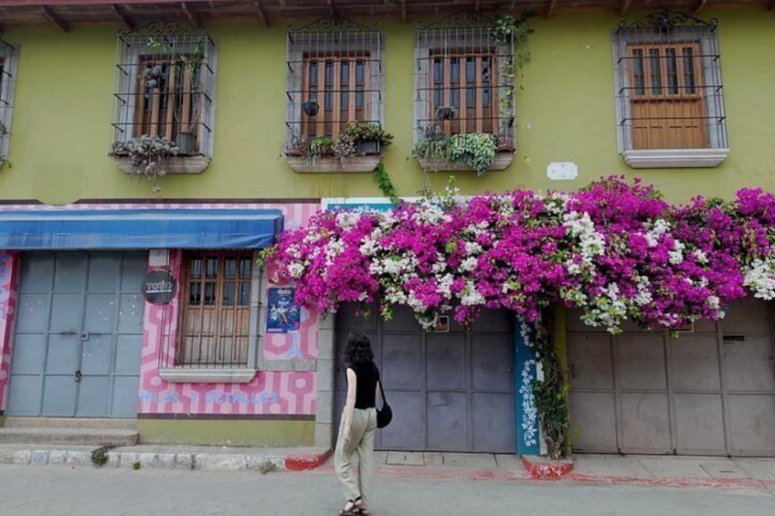 Una calle con un edificio decorado con flores rosadas y blancas, una persona caminando y un toldo azul. 4