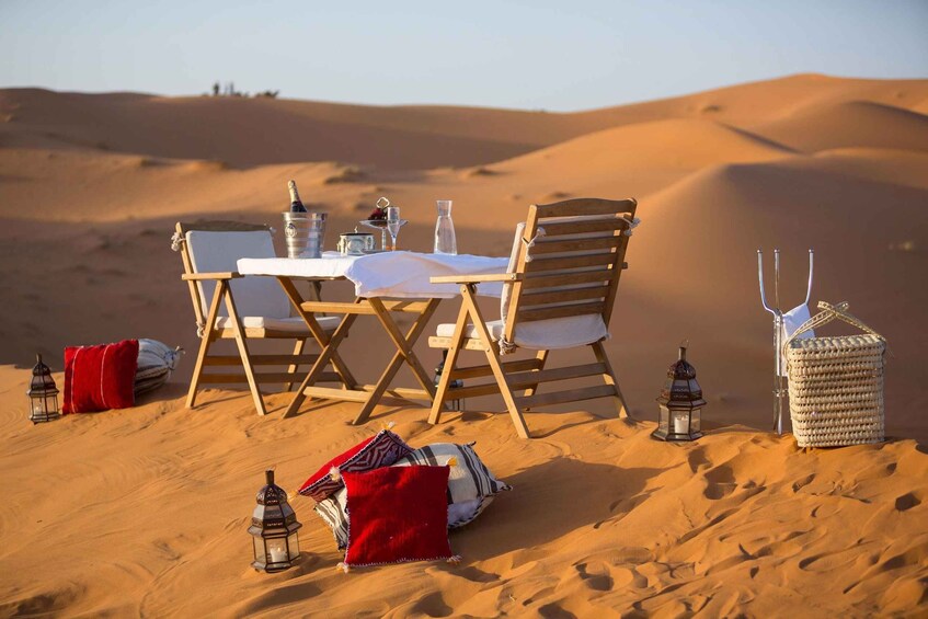 A desert picnic setup with a table, chairs, lanterns, and a woven basket. 2