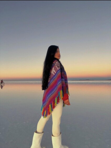 A person standing on a reflective surface with a colorful blanket, wearing white boots, and looking at a sunset over a calm body of water. 3
