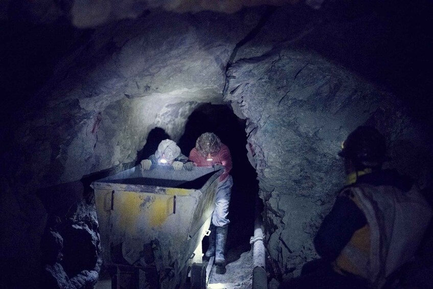 A miner in a tunnel with a headlamp, a cart, and another person in the background. 5