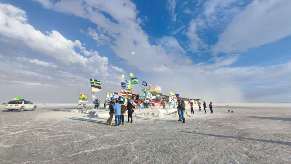 A desert landscape with a clear sky, people standing near a row of flags, and a vehicle in the distance. 5