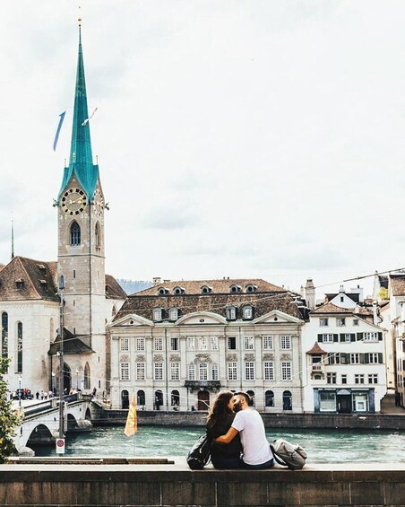 A couple kissing by a river with a clock tower and historic buildings in the background. 5