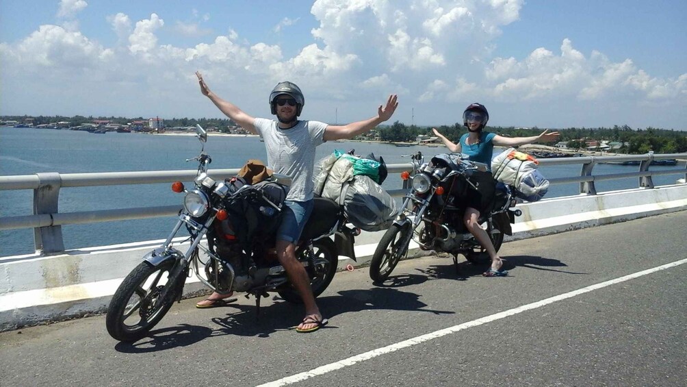 Two people on motorcycles on a bridge with arms raised, overlooking a body of water and distant buildings. 3