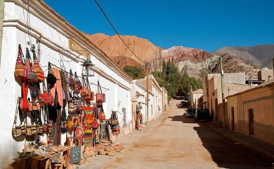 A narrow street with colorful bags hanging on a wall, a sign reading "REGIS", and a mountainous backdrop. 2