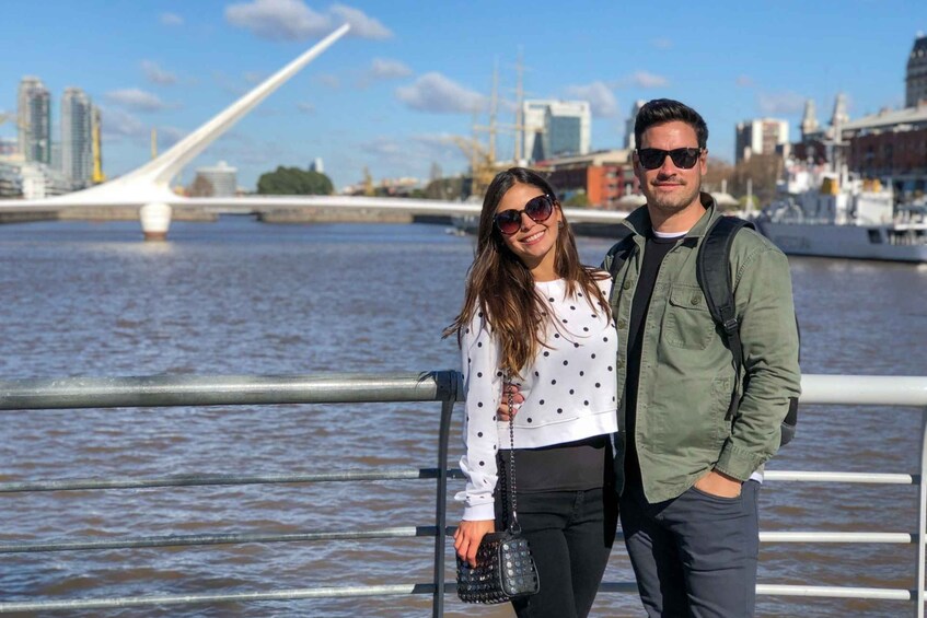 Una pareja de pie junto a la orilla del agua, con un puente moderno y edificios de la ciudad al fondo. 4