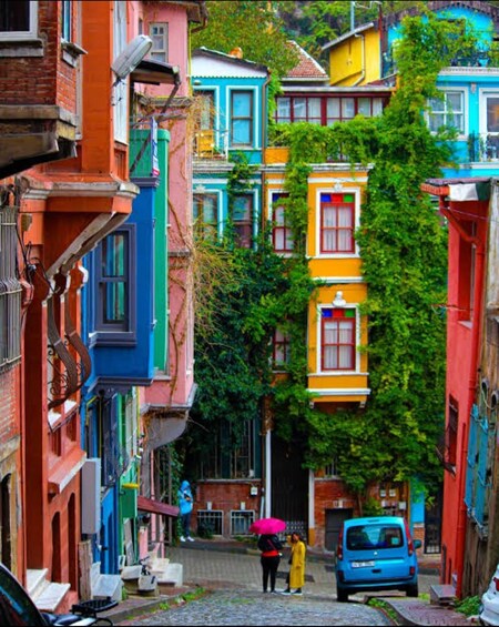 A narrow street lined with colorful buildings, a person with a pink umbrella, and a blue car parked on the right. 1