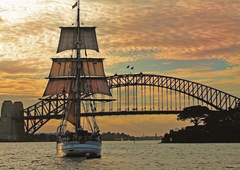 A sailing ship with sails unfurled, crossing the Sydney Harbour Bridge at sunset. 2