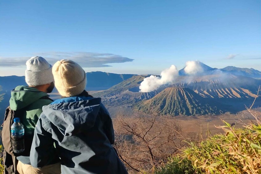 Two people wearing winter hats and jackets, overlooking a mountainous landscape with a volcano emitting smoke. 3