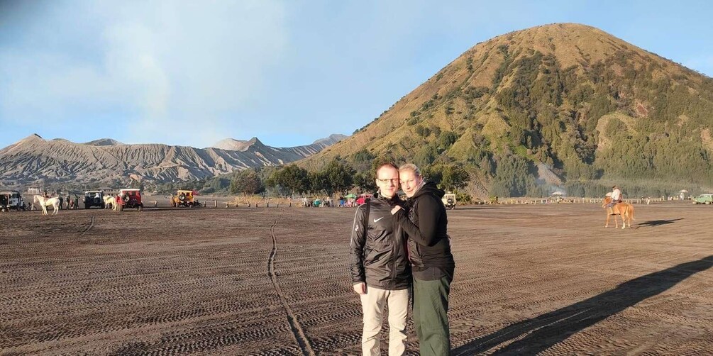 A man and woman standing in a dirt field with horses and vehicles in the background, and a mountain in the distance. 4