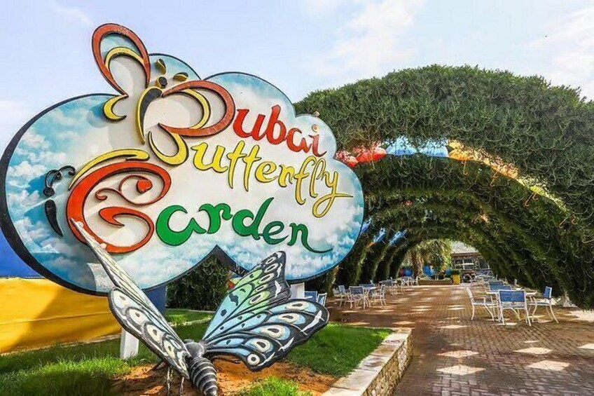 A sign for the Dubai Butterfly Garden with a butterfly design, surrounded by greenery and a paved walkway. 2