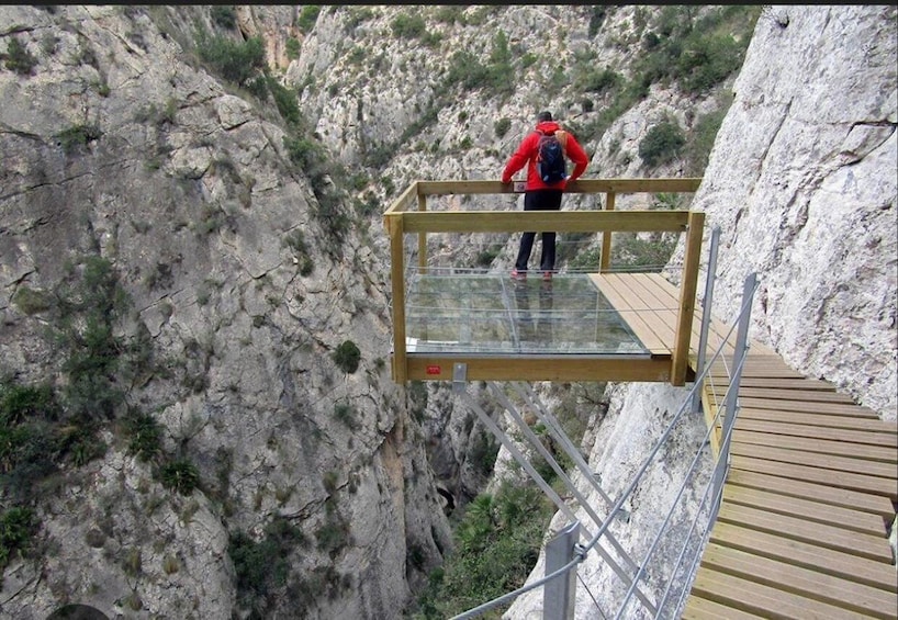 A person standing on a glass bridge with a wooden walkway, overlooking a rocky canyon. 1