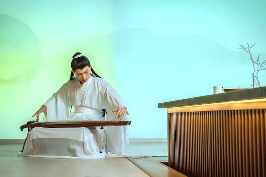 A person in traditional attire playing a stringed instrument, seated on the floor with a wooden table in front. 4