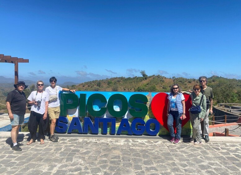 A group of people posing in front of a sign that reads "PICOS SANTAGO" with a mountainous landscape in the background. 1