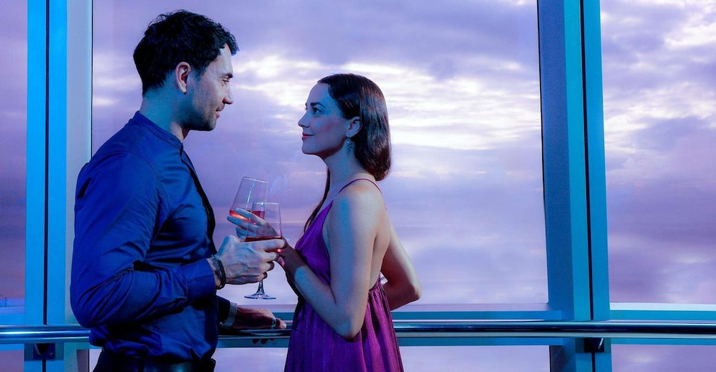 A couple holding wine glasses in front of a window with a view of the sky. 4