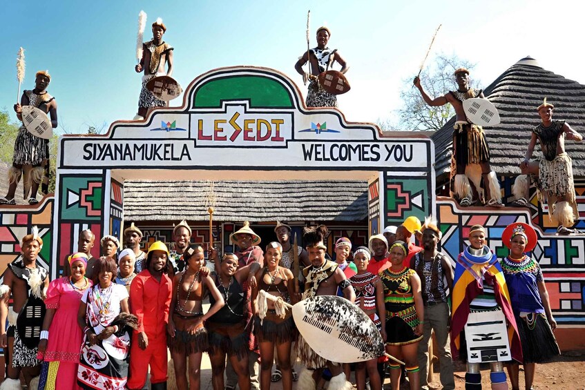 A group of people in traditional attire at a cultural site with a sign reading 'LESEEDI' and 'SIYANAMUKELA WELCOMES YOU'. 1