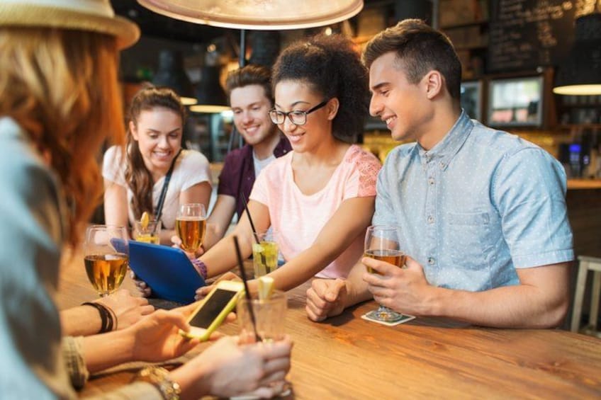 A group of people sitting around a wooden table in a bar, holding drinks and a deck of cards. 2