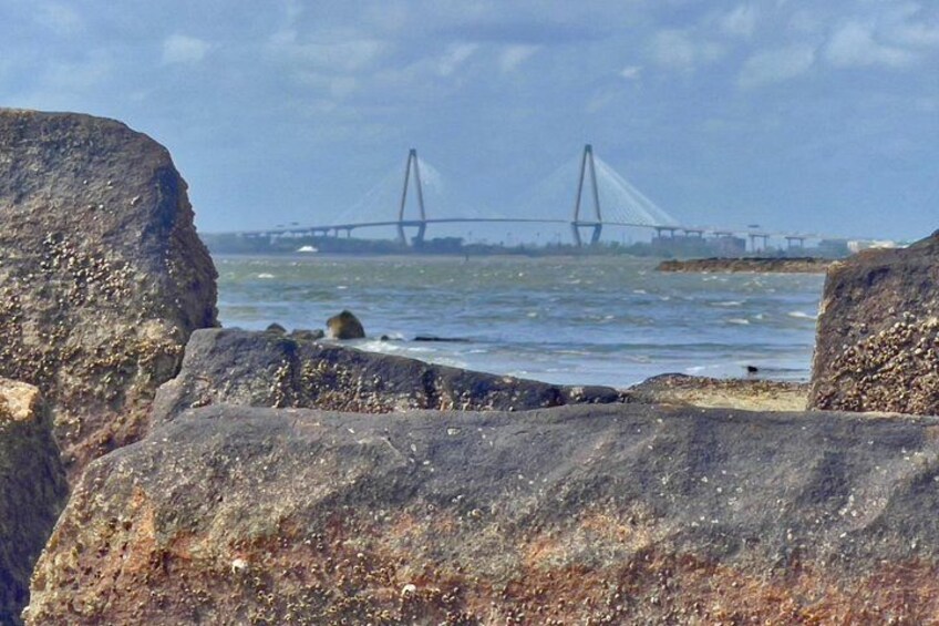A rocky shoreline with large boulders, calm water, and a bridge in the distance. 4