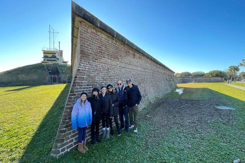 A group of people standing in front of a brick wall with a grassy area and a hill in the background. 3