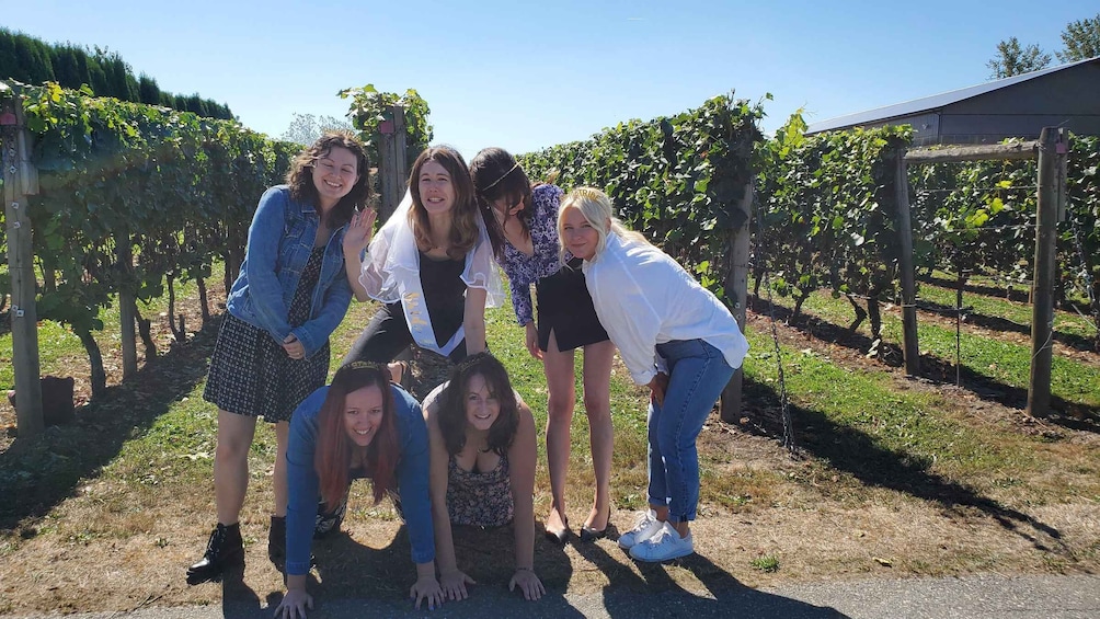 A group of people posing in a vineyard with rows of grapevines and a building in the background. 1