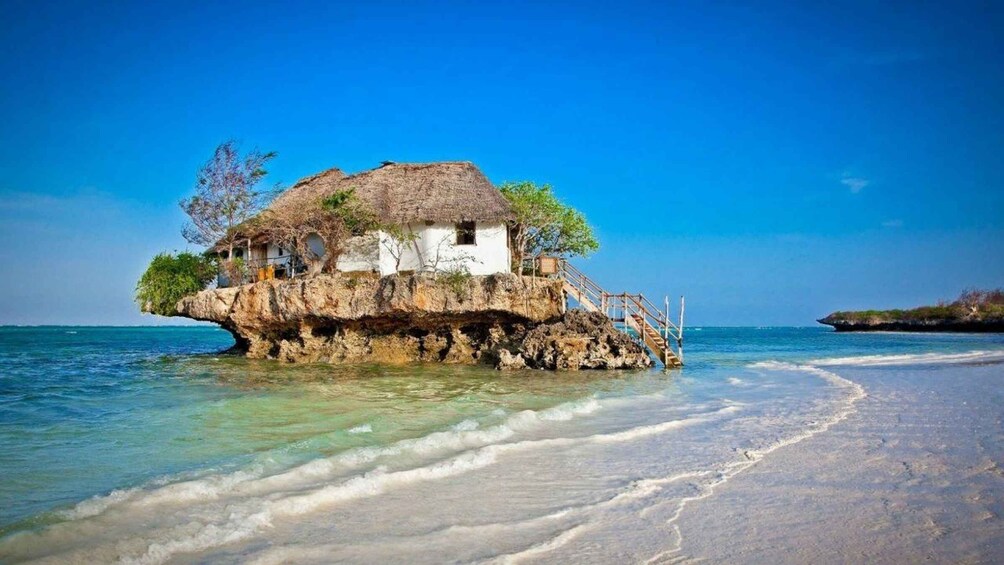 A thatched-roof house on a rocky outcrop with a wooden staircase leading to it, overlooking a beach. 4