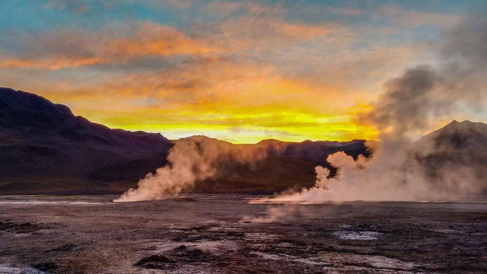A geothermal area with steam vents, a dry lakebed, and mountains in the background during sunset. 1