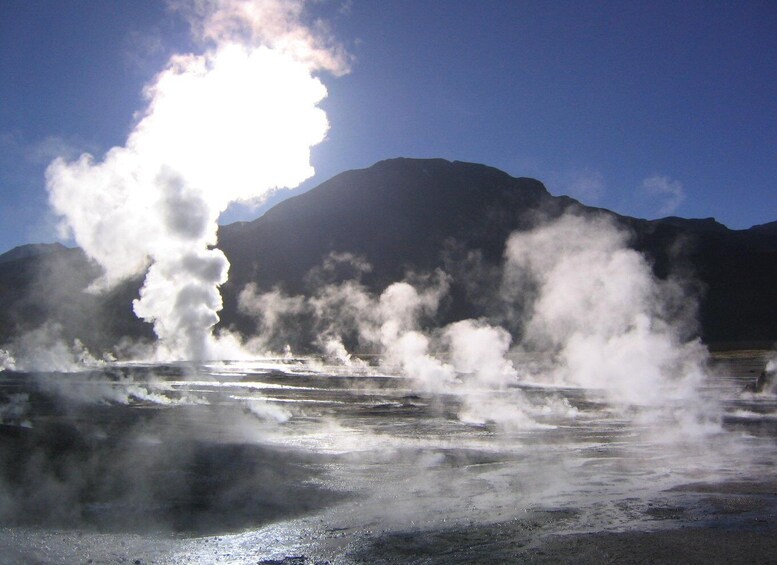 A geyser erupting with steam against a backdrop of mountains and a clear sky. 2